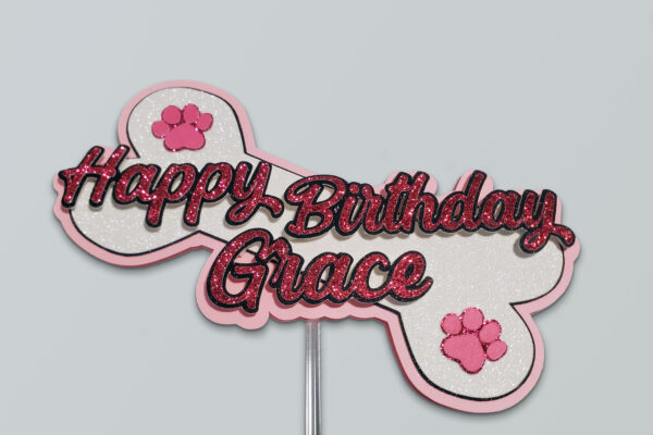 Bone-shaped pink glitter birthday cake topper with the words "Happy Birthday Grace" and two pink paw prints.