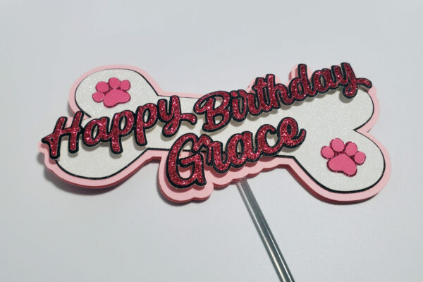 Angled view of pink and white bone-shaped birthday topper for Grace, with glittery text and paw decorations.