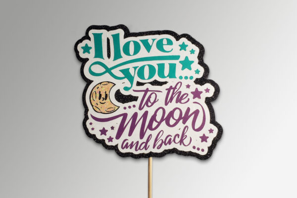Cake topper with teal and purple text that reads ‘I Love You to the Moon and Back,’ accented by glitter, stars, and a smiling moon.