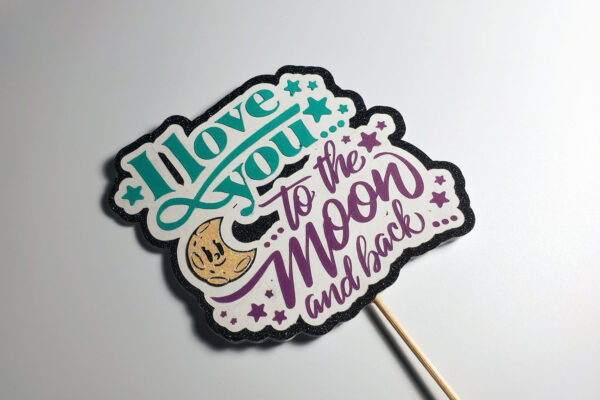 Angled shot of glitter cake topper with text ‘I Love You to the Moon and Back,’ moon graphic, and teal stars.