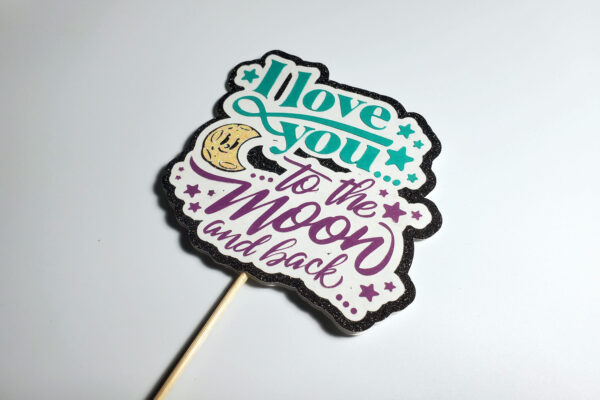 Side view of romantic topper with the phrase ‘I Love You to the Moon and Back’ in teal and purple with black glitter border.