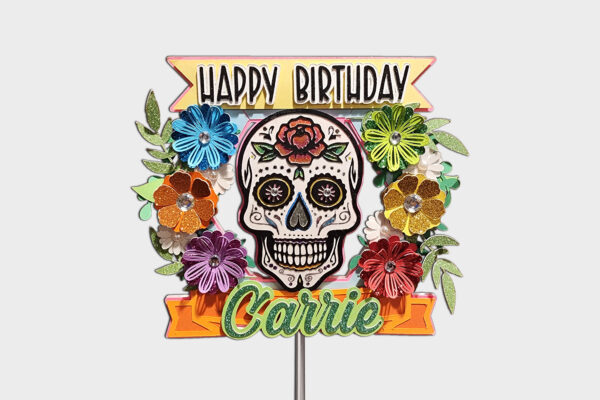 Main shot of a Dia De Los Muertos–themed cake topper featuring a sugar skull at its center, surrounded by layered paper flowers in vivid hues like blue, orange, and green. A ‘Happy Birthday’ banner tops the design, with a customizable name banner below reading ‘Carrie.’ The topper measures 5.5 inches wide and is supported by a clear acrylic stick. Ideal for celebratory events honoring Mexican cultural traditions.