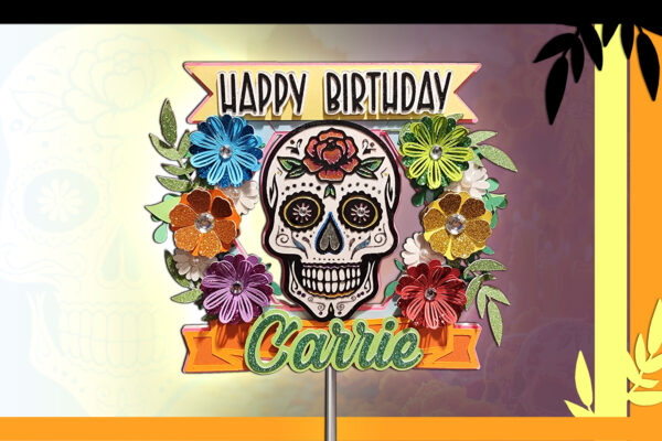 Front-facing view of a Día De Los Muertos sugar skull cake topper adorned with bright paper flowers, glitter accents, and a 'Happy Birthday' banner. The skull has classic Day of the Dead designs with a rose on its forehead. A customizable name banner labeled ‘Carrie’ is at the bottom, and everything is supported by a clear acrylic stick extending several inches below the design.