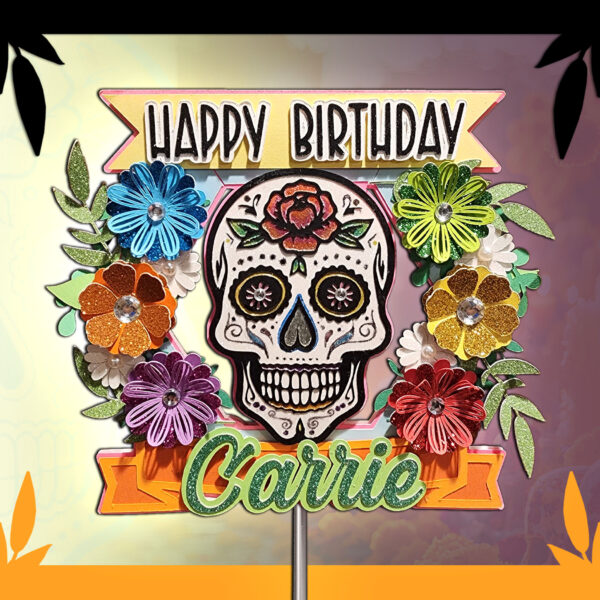 Close-up of a Dia De Los Muertos sugar skull cake topper showcasing vibrant, layered flowers with gem centers, a bold skull design, and glittery text reading ‘Happy Birthday’ and ‘Carrie.’ The beautiful paper construction features colorful leaves and a transparent backing, plus a clear acrylic stick for cake placement. Perfect for Day of the Dead–inspired parties or Mexican-themed celebrations.